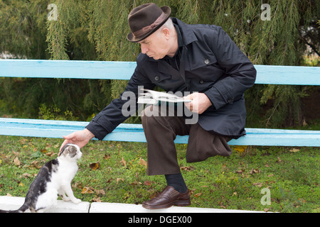 Älteren deaktiviert Mann mit einem Bein amputiert sitzen auf einer Parkbank mit seinem Buch bis hinunter und streicheln einer Katze Stockfoto