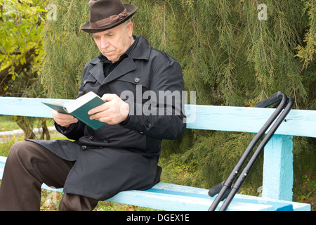 Ältere Menschen deaktiviert Herrn im freien lesen in einen Hut und warmen Mantel auf eine hölzerne Parkbank mit seinen Krücken neben ihm saß. Stockfoto