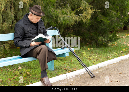 Senior Amputierte genießen sein Buch in der Sonne sitzen auf einer Parkbank mit seinen Krücken neben ihm in einem Hut und warmen Mantel Stockfoto