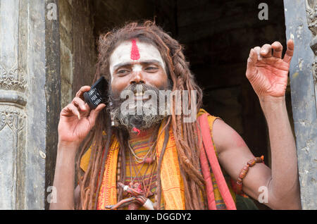 Sadhu, heiliger Mann, mit seinem Handy gegenüber dem brennenden Ghats, Kathmandu, Kathmandu District, Bagmati Zone, Nepal Stockfoto