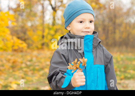 Niedlicher glücklicher kleiner Junge im herbstlichen Wald stehen lächelnd in seine warme gestrickte Beanie Mütze und Jacke mit bunten Blätter in der Hand. Stockfoto