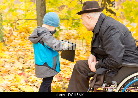 Kleiner Junge zeigt seine behinderte Großvater, die an den Rollstuhl gefesselt ist, durch eine Beinamputation etwas auf seinem Tablettcomputer wie die beiden genießen einen Tag in einem bunten Herbst Park. Stockfoto