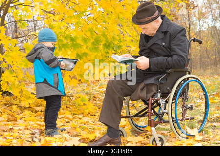 Kleiner Junge mit seinem behinderten Großvater im Freien in einer bunten fallen Garten mit den alten Mann sitzen im Rollstuhl lesen, während sein Enkel auf einem Tabletcomputer spielt. Stockfoto