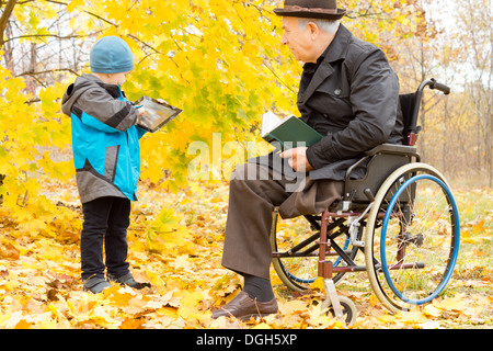 Behinderten älteren Mann mit einem Bein amputiert sitzen in einem warmen Mantel und Hut in seinem Rollstuhl in einem bunten Herbst Park beobachten seines Enkels auf seinem Tablettcomputer zu spielen. Stockfoto