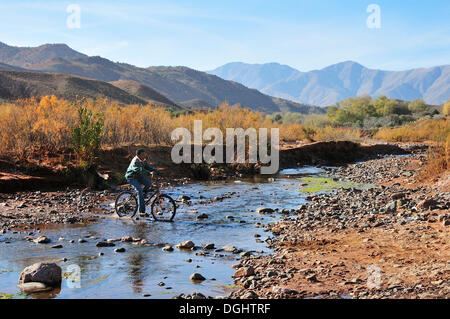 Junge reitet auf seinem Fahrrad durch einen kleinen Fluss in einer kargen Landschaft, Souss-Massa-Draâ, Hoher Atlas, Marokko Stockfoto