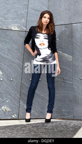 Junge Frau trägt ein Shirt mit einem Porträt-Motiv und Jeans posiert vor einer grauen Wand Stockfoto