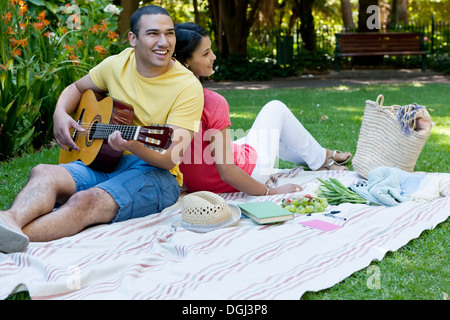 Junges Paar sitzt auf der Picknickdecke, Mann spielt Gitarre Stockfoto