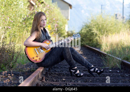 Junge Frau trägt eine Lederweste und Jeans posiert mit einer e-Gitarre auf einem Gleis Stockfoto