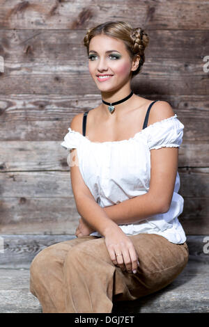 Junge Frau trägt ein weißes Dirndl Top und traditionelle Lederhosen sitzt auf einer Holzbank, Dirndl-Look Stockfoto