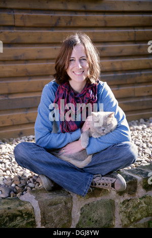 Junge Frau mit einer grauen Katze auf dem Schoß auf einer Mauer sitzend. Stockfoto