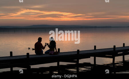 Silhouette eines Paares in der Dämmerung sitzen auf einem hölzernen Pier auf der See Toasten mit Rotweingläser, Steinhude am Meer Stockfoto