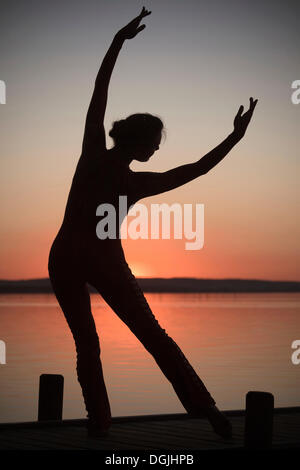 Frau mit Hintergrundbeleuchtung tun eine Yoga-Pose auf einem hölzernen Pier an einem See vor der untergehenden Sonne, Steinhude am Meer Stockfoto