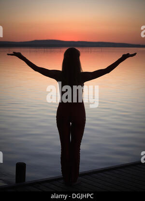 Frau mit Hintergrundbeleuchtung tun eine Yoga-Pose auf einem Holzsteg am See nach Sonnenuntergang, Steinhude am Meer, Niedersachsen Stockfoto