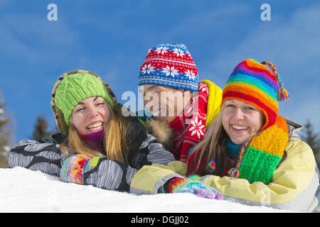 Junge asiatische Mann und zwei junge europäische Frauen Lächeln während liegen auf einem Hügel von Schnee und tragen bunte Winterkleidung Stockfoto