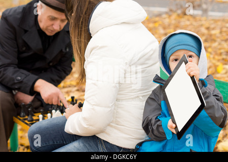 Kleiner Junge gebündelt, in warme Kleidung gegen die kalten Herbstwetter sitzt auf einer Parkbank mit einem Tabletcomputer zu spielen, während seine Mutter und Großvater Schach mit ihm spielen. Stockfoto