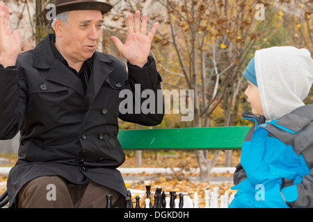 Niedliche kleine Junge Konfrontation mit seinem Großvater, die spielerisch seine Unschuld mit seinen Händen in die Luft erhoben protestieren ist, wie sie eine Partie Schach auf einer Parkbank spielen. Stockfoto