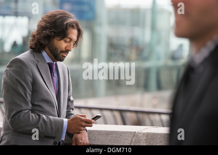 Zwei Männer in Business-Anzügen, auf einer Terrasse mit einem Geländer. Eine Überprüfung sein Telefon. Stockfoto