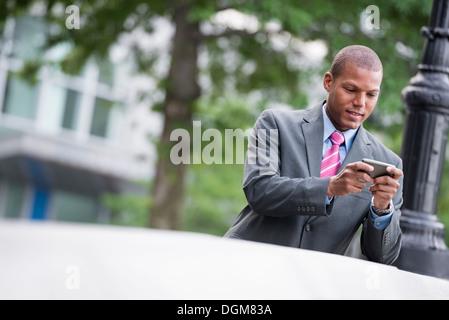 Ein junger Mann in einem Business-Anzug, ein hellblaues Hemd mit roter Krawatte. In einer New-York-City-Straße. Verwenden ein smart Phone. Stockfoto