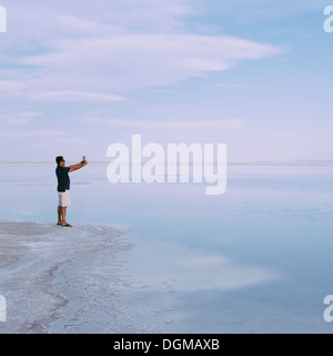 Ein Mann steht am Rand des überfluteten Bonneville Salt Flats in der Abenddämmerung, Fotografieren mit einem Tablet-Gerät in der Nähe von Wendover. Stockfoto