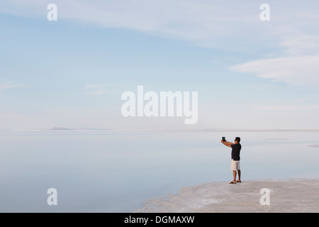 Ein Mann steht am Rand des überfluteten Bonneville Salt Flats in der Abenddämmerung, Fotografieren mit einem Tablet-Gerät in der Nähe von Wendover. Stockfoto