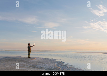 Ein Mann steht am Rand des überfluteten Bonneville Salt Flats in der Abenddämmerung, Fotografieren mit einem Tablet-Gerät. Stockfoto