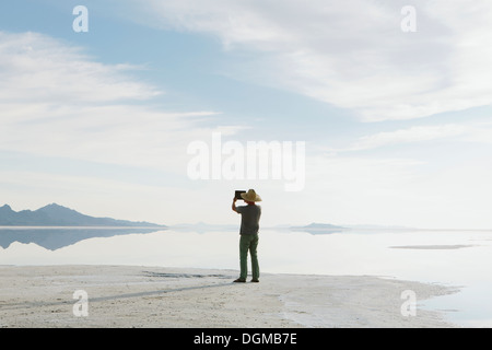 Ein Mann steht am Rand des überfluteten Bonneville Salt Flats in der Abenddämmerung, Fotografieren mit einem Tablet-Gerät. Stockfoto