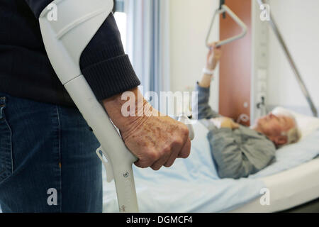 Ältere Frau, die auf eine Krücke vor einem älteren Mann in einem Krankenhausbett liegen Stockfoto