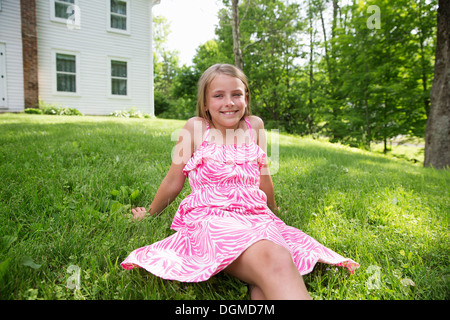 Ein junges Mädchen in einem rosa gemusterten Sommerkleid sitzen auf dem Rasen unter den Bäumen in einen Bauerngarten. Stockfoto