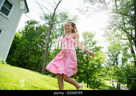 Ein junges Mädchen in einem rosa gemusterten Sommerkleid quer über den Rasen unter den Bäumen in einen Bauerngarten. Stockfoto