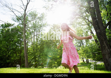 Ein junges Mädchen in einem rosa gemusterten Sommerkleid quer über den Rasen unter den Bäumen in einen Bauerngarten. Stockfoto