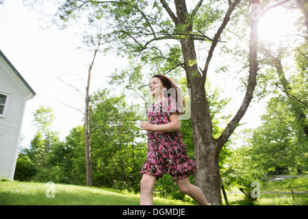 Ein junges Mädchen in einem gemusterten Sommerkleid, quer über den Rasen im Schatten der Bäume in einem Bauerngarten. Stockfoto