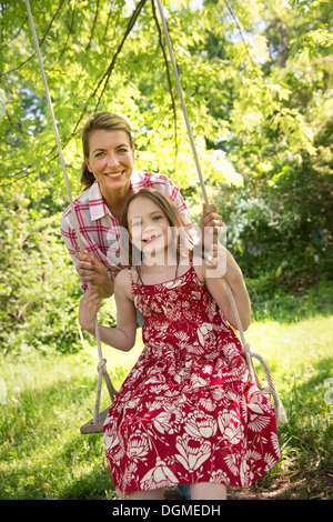 Sommer. Eine Mädchen in einem Sommerkleid auf einer Schaukel hing an einem Ast. Eine reife Frau hinter ihr. Stockfoto