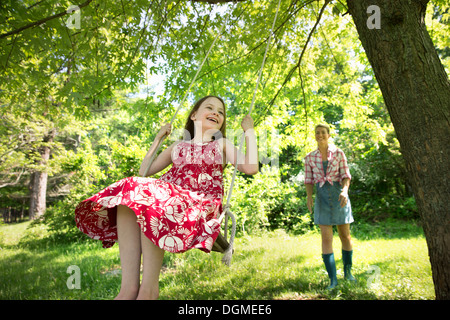 Sommer. Ein Mädchen in einem Sommerkleid auf einer Schaukel unter einem grünen Baum. Eine Frau, die hinter ihr stehen. Stockfoto