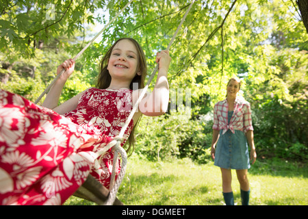 Sommer. Ein Mädchen in einem Sommerkleid auf einer Schaukel unter einem grünen Baum. Eine Frau, die hinter ihr stehen. Stockfoto