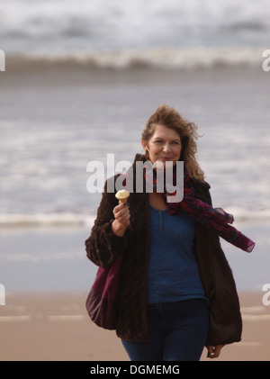 Mittlere gealterte Frau zu Fuß am Strand mit einem Eis im Winter, Bude, Cornwall, UK Stockfoto