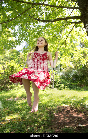 Sommer. Ein Mädchen in einem Sommerkleid auf einer Schaukel schwingen von Ast eines belaubten Baumes. Stockfoto