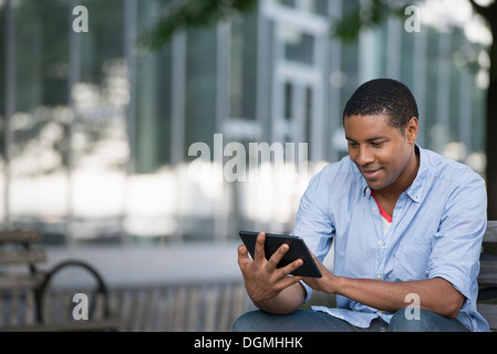 Sommer in der Stadt. Menschen im Freien, reibungslose Kommunikation unterwegs. Ein Mann sitzt auf einer Bank mit einem digitalen Tablet. Stockfoto