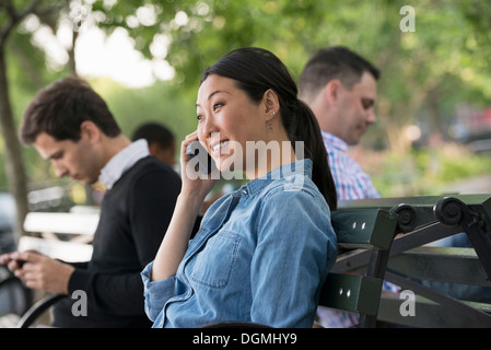 Sommer in der Stadt. Eine Frau und drei Männer sitzen im Park, jede auf ihrem eigenen Handy oder verwenden eines Tablet. Stockfoto