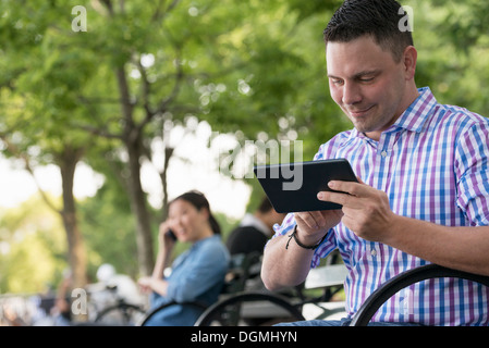 Sommer. Eine Frau und zwei Männer sitzen im Park, jede auf ihrem eigenen Handy oder verwenden eines Tablet. Stockfoto