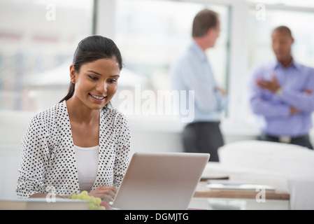 Büroalltag. Eine Frau sitzt an einem Schreibtisch mit einem Laptopcomputer. Zwei Männer im Hintergrund. Stockfoto