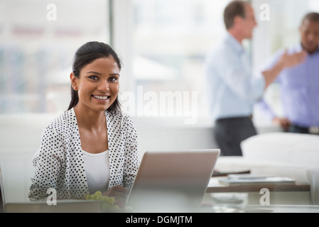 Büroalltag. Eine Frau sitzt an einem Schreibtisch mit einem Laptopcomputer. Zwei Männer im Hintergrund. Stockfoto