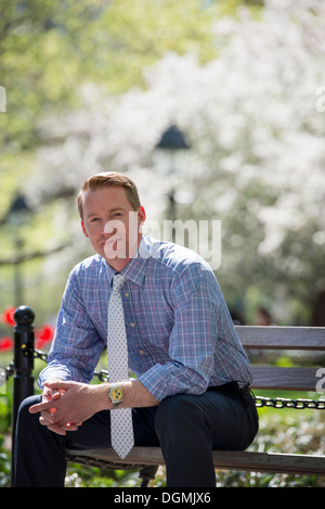 Ein Geschäftsmann in einem Hemd mit Frack, sitzen auf einer Parkbank im Schatten eines Baumes mit Blüte. Stockfoto