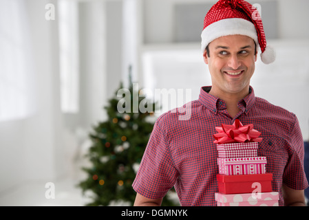 Büroeinrichtung. Weihnachten zu feiern. Ein Mann in eine Weihnachtsmütze hält einen Stapel von Geschenken. Geschmückter Weihnachtsbaum. Stockfoto