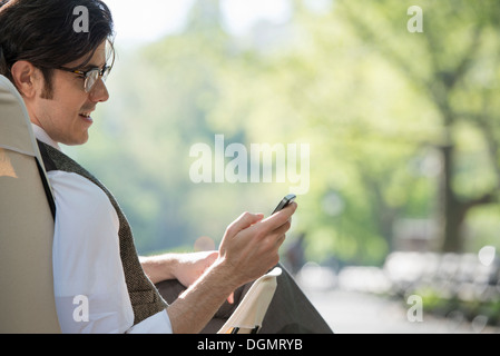 Leben in der Stadt. Ein Mann sitzt in einer Leinwand Campingstuhl in den Park, Blick auf sein Handy. Stockfoto