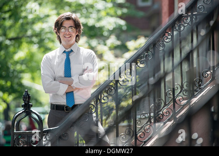 Stadt. Ein junger Mann in w weißes Hemd und eine blaue Krawatte, stehend mit Armen gefaltet außerhalb eines Stadthauses, auf den Stufen. Stockfoto