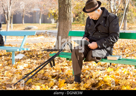 Senior deaktiviert Man mit einem Bein auf einem Tablet-pc im Internet surfen, wie er in einen warmen Mantel und Hut genießen einen sonnigen Herbsttag im Freien im Park sitzt. Stockfoto