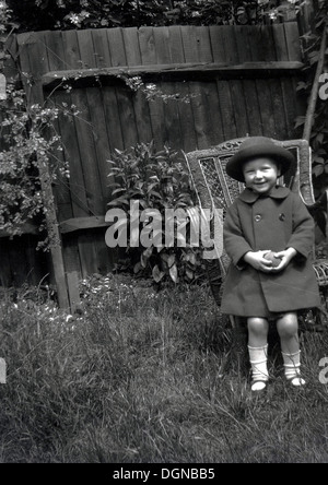 Historische Aufnahme aus den 1930er Jahren eines elegant gekleideten Mädchens tragen Mantel und Hut steht in einem Garten hinter dem Haus. Stockfoto