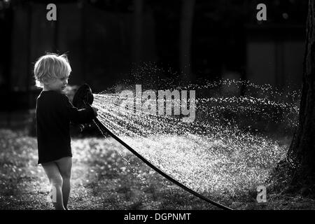 Kleiner Junge spielt mit einem Wasserschlauch im Garten, mit Hintergrundbeleuchtung, Berlin, Berlin, Deutschland Stockfoto