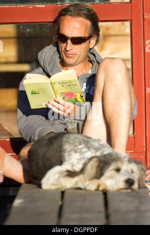 Mann auf einem Pier in der Sonne sitzen und lesen ein Buch mit seinem Hund schlafen vor ihm, Weißensee, Kärnten, Österreich Stockfoto