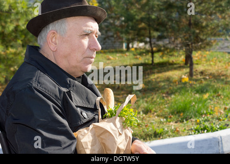 Älterer nachdenklicher Mann in Mantel und Hut mit einer Tasche von Lebensmitteln auf seinem Schoß in der Sonne sitzt hautnah Seite Ansicht Porträt. Stockfoto
